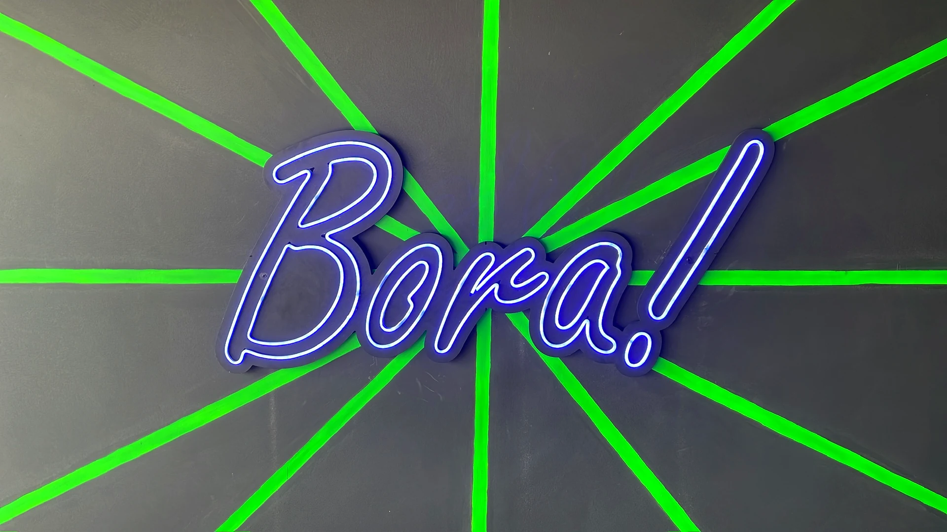 Letreiro luminoso Bora! em uma parede cinza com faixas verdes